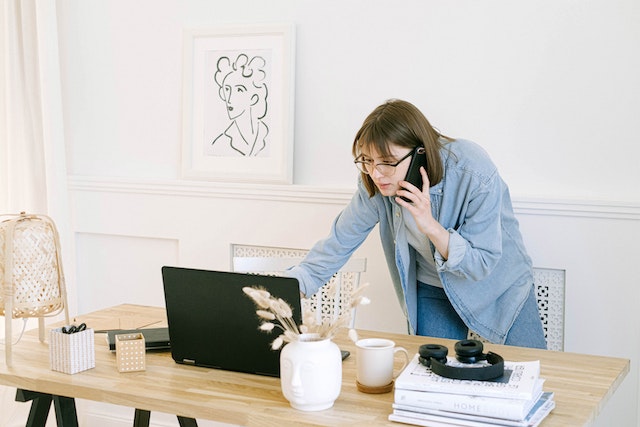 a person standing at a desk on the phone, looking at a laptop